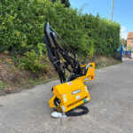 Yellow construction machine with a hydraulic arm on a street, next to a tall green hedge and a red-brick building in the background, under a blue sky