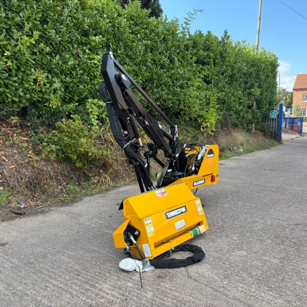 Yellow construction machine with a hydraulic arm on a street, next to a tall green hedge and a red-brick building in the background, under a blue sky