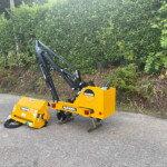 Yellow McConnel construction attachment with hydraulic arms on a black frame, parked in a driveway beside a tall green hedge.