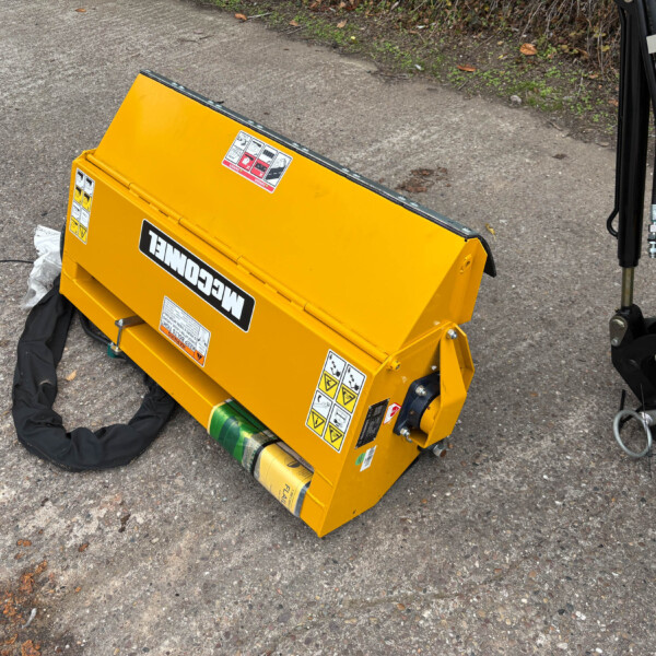 Yellow construction attachment with warning stickers lying on gravel, hoses and a black hydraulic post nearby—appears to be a piece of heavy equipment.