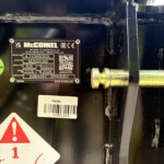 Black metal machinery with an identification plate reading 'McCONNEL' and model/serial details; a bright green 'Passed Inspection' sticker nearby; a white barcode label and partial red warning sign are visible.