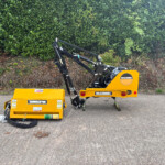Yellow McConnel construction attachment with hydraulic arms parked on asphalt, hedge in the background