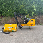 Yellow McConnel agricultural machine with hydraulic arms parked on a road in front of a tall green hedge at the edge of a field.