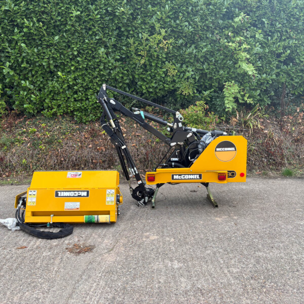 Yellow McConnel agricultural machine with hydraulic arms parked on a road in front of a tall green hedge at the edge of a field.