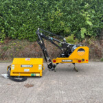 Yellow McConnel construction machine with hydraulic arms resting on a pavement, dense green hedge in the background.