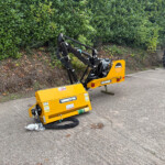 Yellow McConnel hydraulic attachment on a trailer on a road, with hydraulic arms raised and a red Manitou tractor visible in the background by a green hedge.