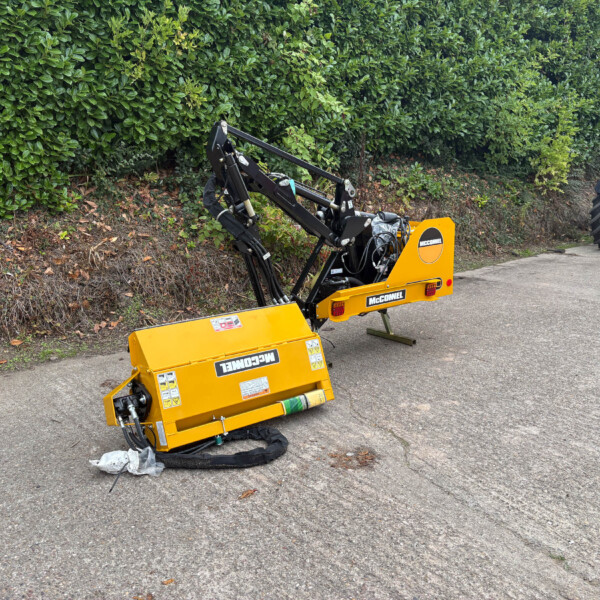Yellow McConnel hydraulic attachment on a trailer on a road, with hydraulic arms raised and a red Manitou tractor visible in the background by a green hedge.