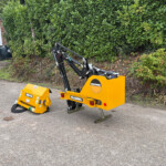 Yellow McConnel hydraulic attachment on a compact construction machine in a paved lot, with a hedge and building in the background.