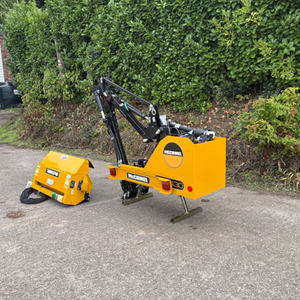 Yellow McConnel hydraulic attachment on a compact construction machine in a paved lot, with a hedge and building in the background.