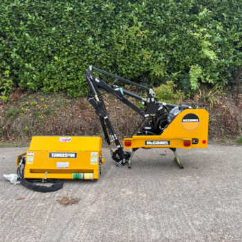 Yellow McConnel construction machine with hydraulic arms resting on a pavement, dense green hedge in the background.