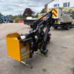 Yellow industrial machine with a hydraulic arm in a parking lot, fronts a row of blue tractors and a building with 'RUSSELL' signage in the background