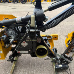 Close-up of yellow industrial machinery with hydraulic cylinders, hoses, and warning stickers visible at a construction site.