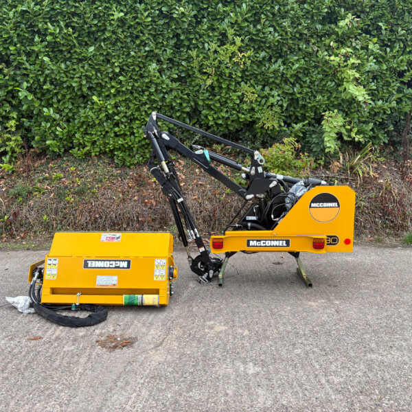 Yellow McConnel construction machine with hydraulic arms resting on a pavement, dense green hedge in the background.