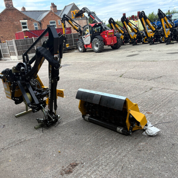 Industrial yard with yellow and black construction machinery; foreground shows a hydraulic lifting arm, background rows of tractors with attachments and a brick house fence behind.