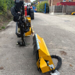 Close-up of a yellow hydraulic construction attachment on a mobile machine parked on a street, with a blue gate and red trailer in the background.
