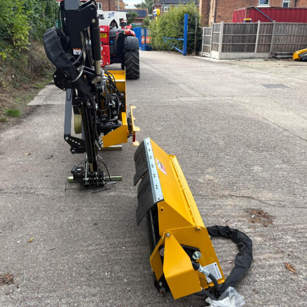 Close-up of a yellow hydraulic construction attachment on a mobile machine parked on a street, with a blue gate and red trailer in the background.