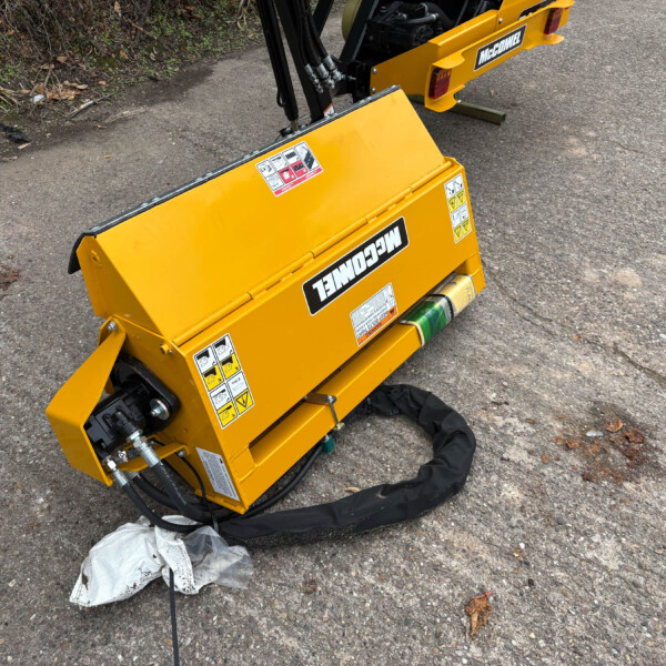 Close-up of a yellow construction attachment on pavement, with warning labels and hydraulic hoses visible.