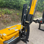 Yellow hydraulic machinery attachment with hoses and levers mounted on a black metal frame, outdoors beside a hedge; safety labels visible.