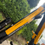 Close-up of a yellow construction machine arm with black hydraulic hoses and bolts, set against a green hedge outdoors.