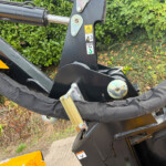 Close-up of a heavy construction machine's hydraulic arm and black metal frame with caution stickers and a 2500 kg load label, outdoors with green shrubbery in the background.