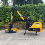 Yellow hedge-trimming machine with an extendable black arm parked on a paved surface, dense green hedge in the background.
