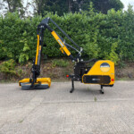 Compact yellow and black hydraulic lifting arm machine parked on a paved surface with a green hedge in the background, McConnel branding visible on the side.