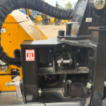 Close-up of a yellow heavy-duty machine with a worker's dark-clad arm, black frame, and safety labels visible.