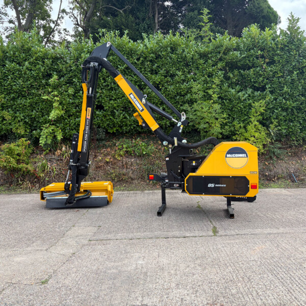 Yellow McConnell 85 Series compact excavator with extended hydraulic arm parked on a paved lot, hedge in background.