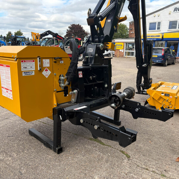 Close-up of a yellow industrial machine with black hydraulic arms in a dealership lot, with blue tractors and a Russell's building in the background.