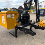 Yellow industrial hydraulic machine on a trailer in a dealership lot, with blue tractors and a Russell storefront in the background.