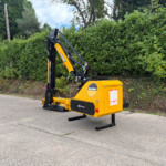Yellow McConnel 85-series hydraulic machine with an extended arm in a parking area beside a dense hedge, ready for work