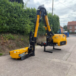Orange‑and‑black construction machine with hydraulic arms on a street beside a tall green hedge, a brick building in the background.