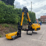 Yellow hydraulic demolition/rigging machine with an articulated arm parked on a street beside a tall hedge, with a red brick building in the background.