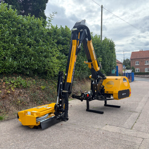 Yellow hydraulic demolition/rigging machine with an articulated arm parked on a street beside a tall hedge, with a red brick building in the background.