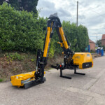 Yellow articulated construction machine with hydraulic arms parked on a street beside a tall hedge and brick buildings in the background.