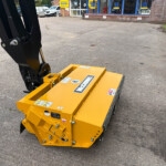 Close-up of a yellow hydraulic attachment with a black arm, parked on asphalt near storefronts in the background.