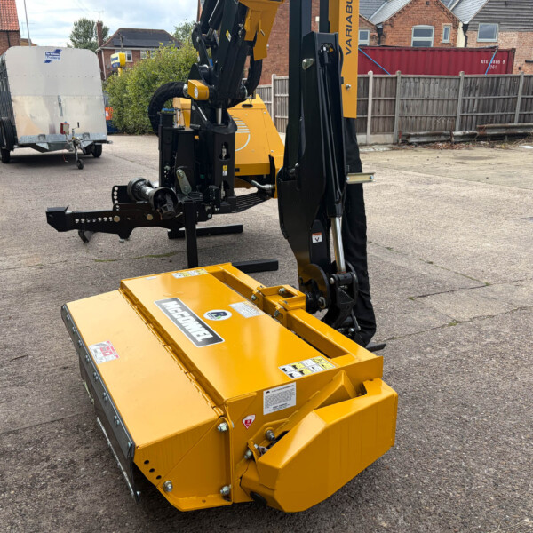 Yellow hydraulic construction machine with black arm and front attachment on a paved area, trailer in the background and brick houses nearby.