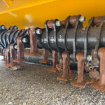 Close-up of rusted metal blades and bolts on the underside of a yellow agricultural machine, with gravel ground beneath.