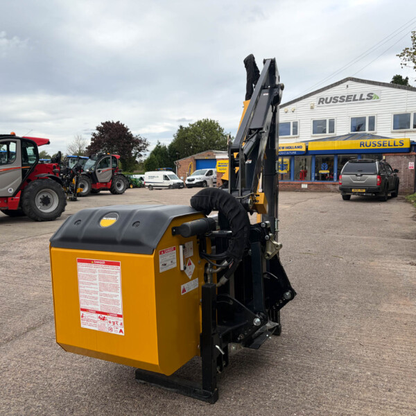 Close-up of a yellow industrial machine with warning stickers in a machinery lot, with red and gray tractors and a Russell's store in the background.