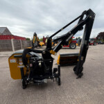 Close-up of a yellow construction machine with a large black lifting arm in a lot, other equipment and brick houses in the background.