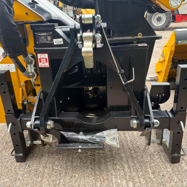Close-up of a yellow construction attachment's hydraulic hitch with safety labels and bolts visible.