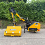 Yellow McConnel stump grinder attachment mounted on a compact tractor, parked on a paved surface with a dense green hedge in the background.