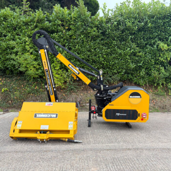 Yellow McConnel landscaping machine with a mower attachment, parked on a paved surface with a green hedge in the background,