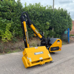 Yellow compact excavator parked on a paved road beside a tall green hedge, with a brick building in the background.