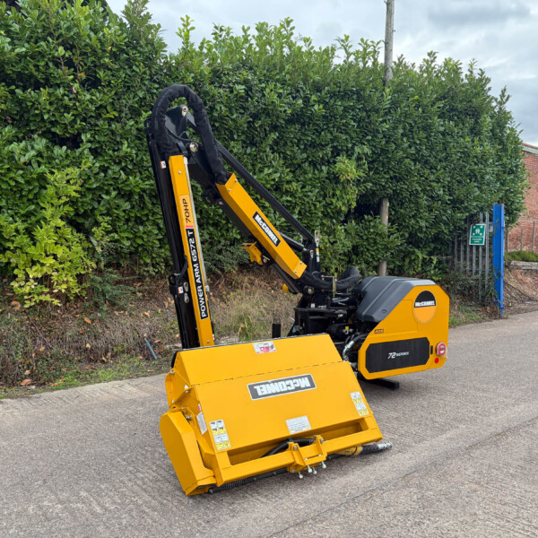 Yellow compact excavator parked on a paved road beside a tall green hedge, with a brick building in the background.