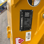 Close-up of a yellow industrial machine showing a black rectangular data plate and safety stickers on the frame (McConnel label, QR code).