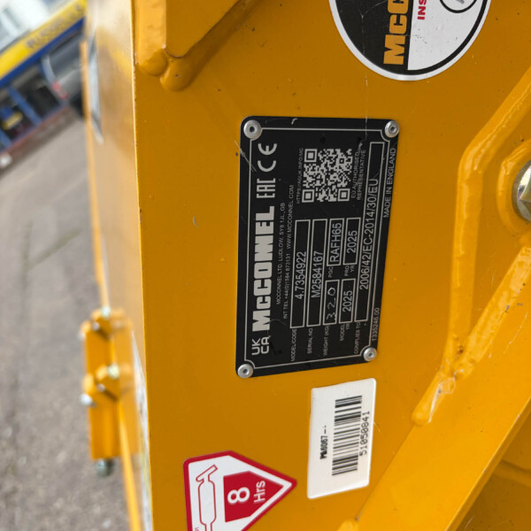 Close-up of a yellow industrial machine showing a black rectangular data plate and safety stickers on the frame (McConnel label, QR code).