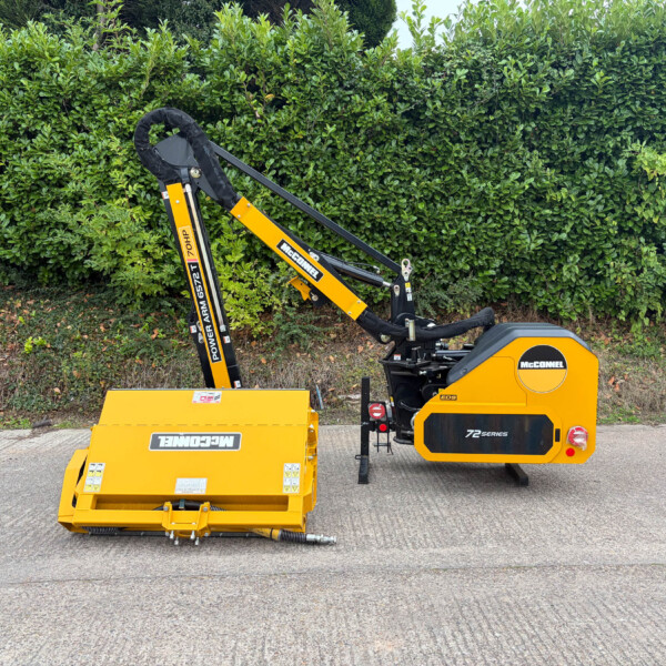 Yellow McConnel stump grinder attachment mounted on a compact tractor, parked on a paved surface with a dense green hedge in the background.
