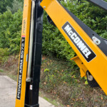 Close-up of a yellow hydraulic excavator arm with a black frame and brand stickers beside a hedge.