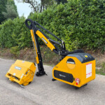Yellow McCONNELL mini excavator with folded arm on a paved surface, adjacent to a hedge and a detached yellow attachment lying on the ground in front.